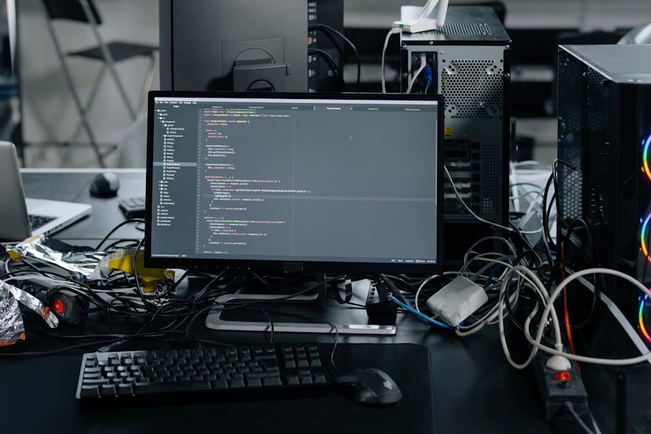 A cluttered office workspace featuring a computer monitor displaying lines of code, surrounded by tangled cables and electronic devices. To the left, there is a laptop and a keyboard, with some crumpl